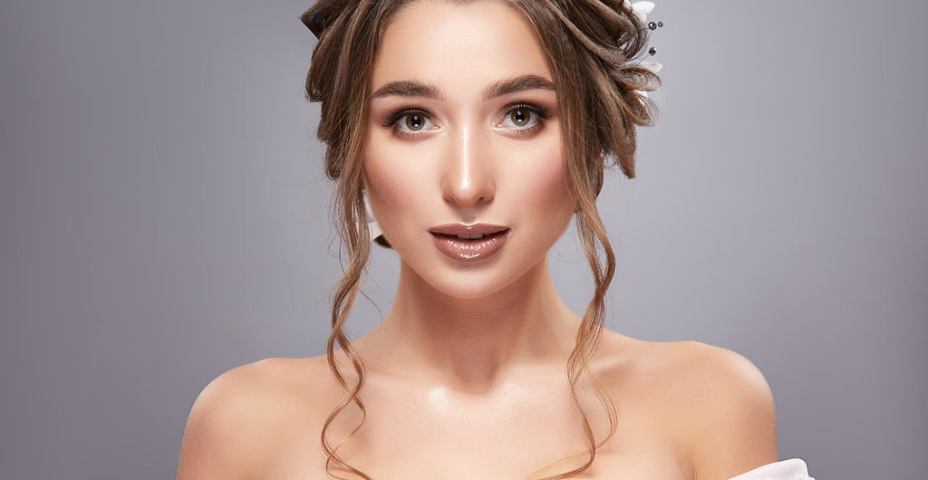 Woman wearing a wedding gown with a full face of bridal makeup and full bridal hair on grey background