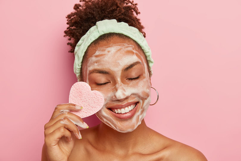 Woman doing skincare in front of pink background with a heart-shaped sponge next to her face