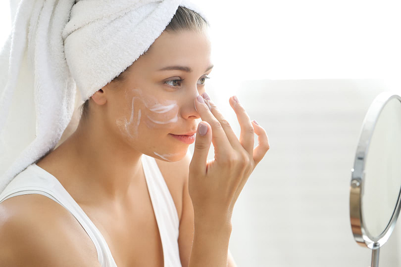 A woman applying moisturizer on her face in front of a mirror